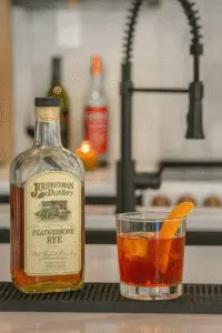 Featherbone Rye Boulevardier at Copper Top Ridge Kitchen Bar Featherbone Rye Boulevardier cocktail served on a black bar mat atop a white marble countertop at Copper Top Ridge, with a bottle of Journeyman Distillery rye whiskey and an orange peel garnish.