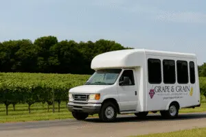 White Grape & Grain winery tour shuttle bus driving past rows of lush green vineyard under a sunny blue sky.