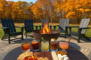 A cozy autumn scene at Copper Top Ridge showing cocktails around the bonfire. Five fall-inspired drinks sit on a table with a gourmet s’mores charcuterie board in the foreground, while a glowing fire pit and Adirondack chairs are surrounded by colorful fall foliage.